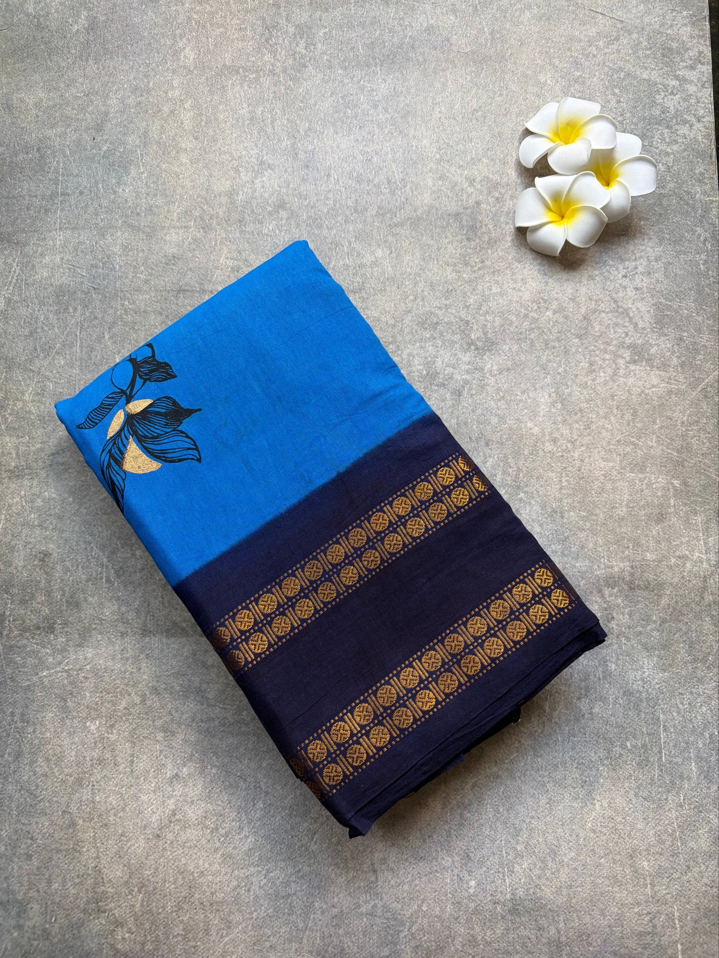 Blue and navy sungudi saree with gold patterns on a gray surface, accompanied by white flowers.