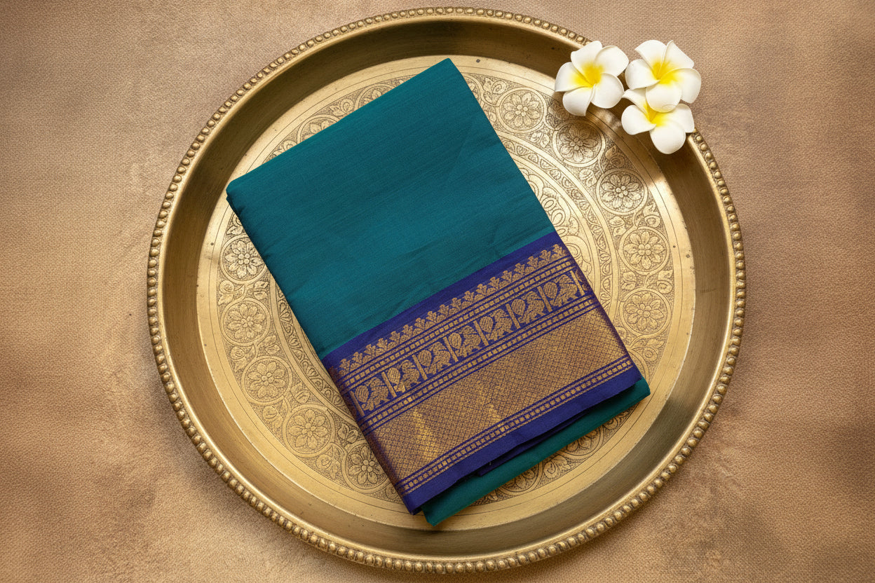 Teal saree with a gold border on a beige surface with white flowers.